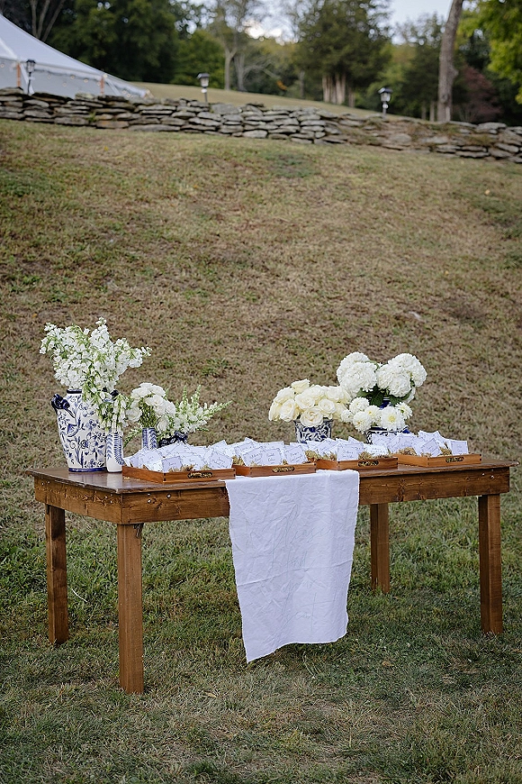 Escort card display with wedding escort cards on a wood table, blue-and-white ceramic vases and white flowers on a lawn by a stone wall
