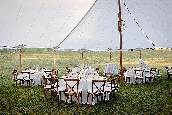 Reception tablescape under an outdoor tent reception with white linens, crossback chairs, tall glass vases, candles, and string lights on a lawn