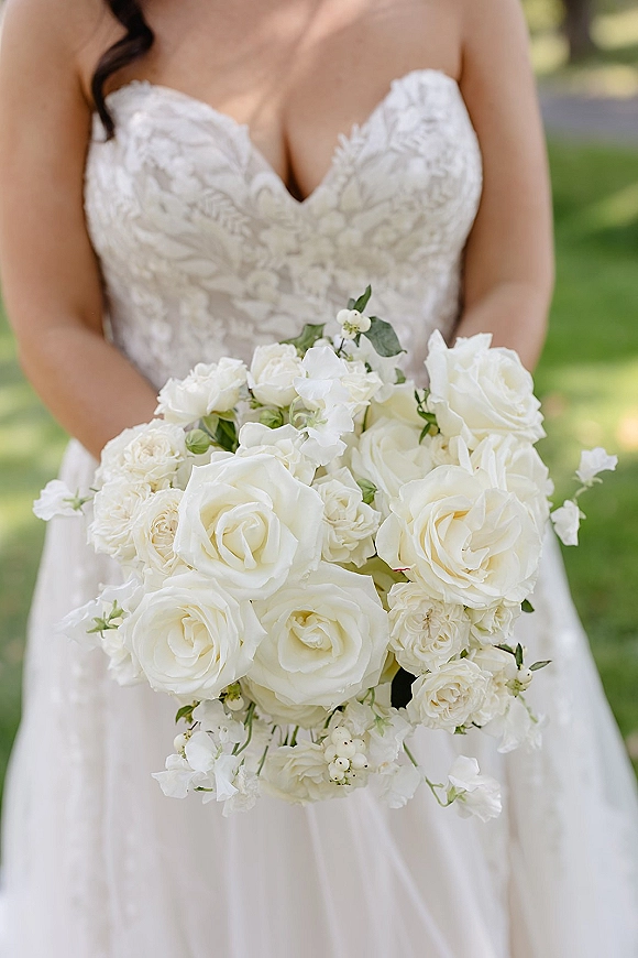 Bridal bouquet of white roses with greenery and sweet pea blooms held against a strapless lace bodice on a sunny lawn with trees