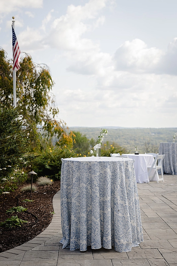 Cocktail table decor with a blue and white tablecloth and bud vase of white flowers on an outdoor patio with hillside views