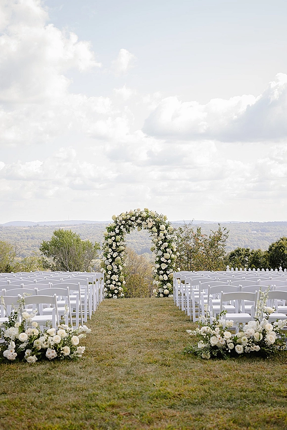 Ceremony setup with an outdoor wedding ceremony floral arch of white roses and greenery, lined with white chairs on a grassy hilltop lawn