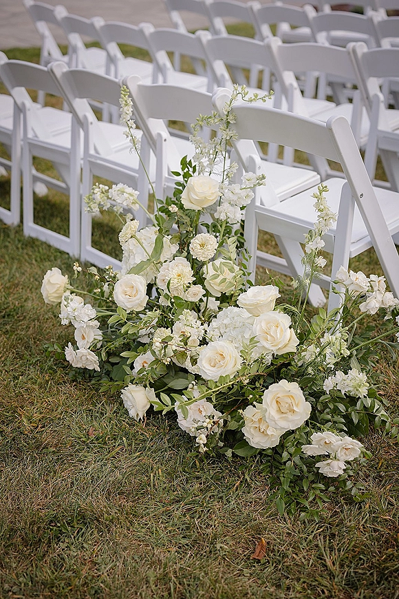 Ceremony aisle flowers with ground floral aisle decor of white roses and greenery lining white folding chairs on a grass lawn outdoors