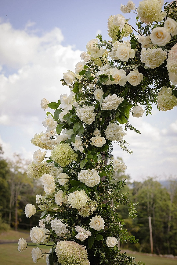 Floral wedding arch with white roses and hydrangeas draped in greenery, set in a meadow under blue sky and clouds