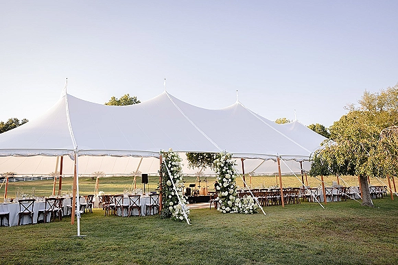 Wedding tent reception with sailcloth tent wedding details, long banquet tables and floral arch entrance on a grassy lawn with trees
