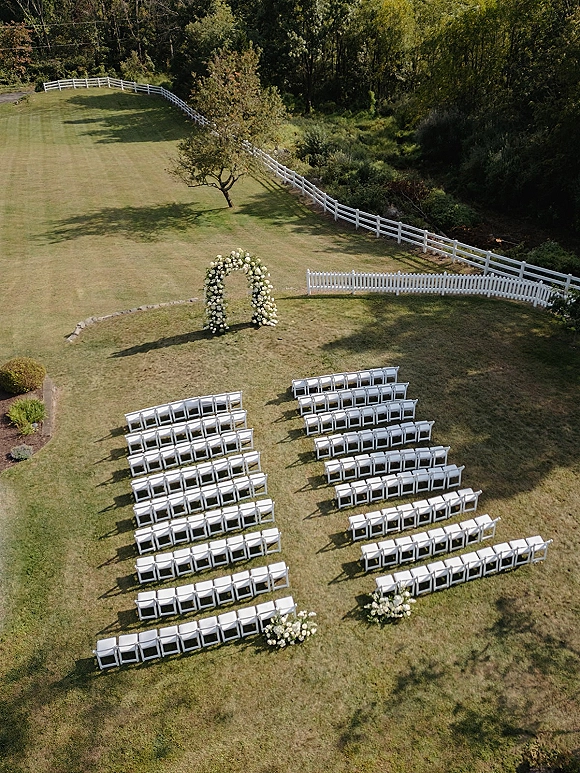 Outdoor ceremony setup with white folding chairs facing a floral ceremony arch of white roses and greenery on a grassy lawn by a white fence