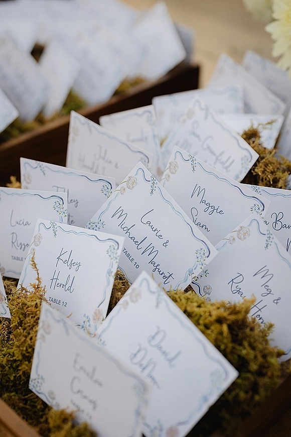 Wedding escort cards with calligraphy names and blue floral borders nestled in moss-filled wooden boxes on a neutral tabletop