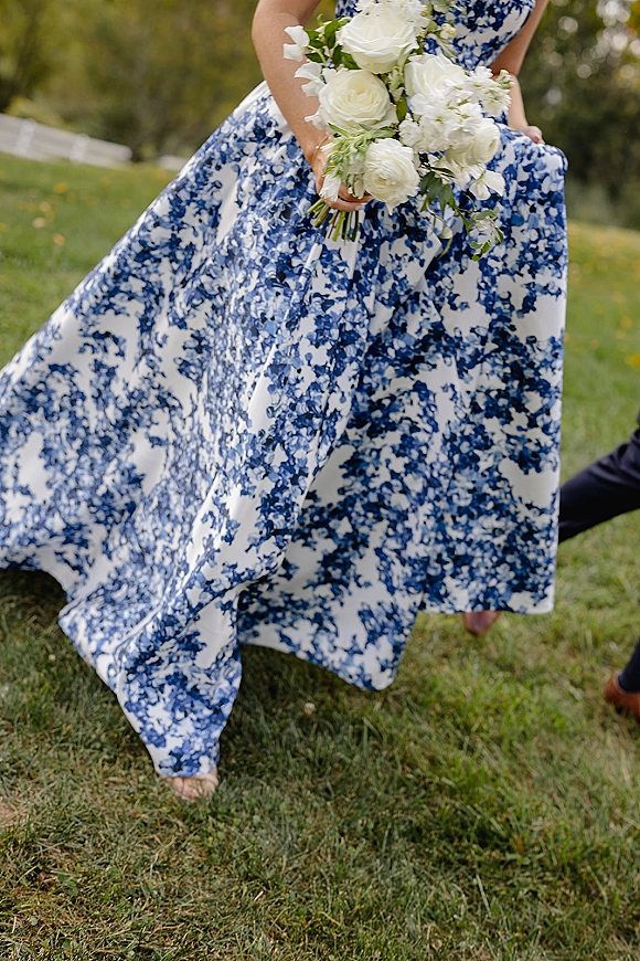 Wedding dress with blue and white floral pattern as the bride walks on a grassy lawn, holding a white rose bouquet with ribbon wrap
