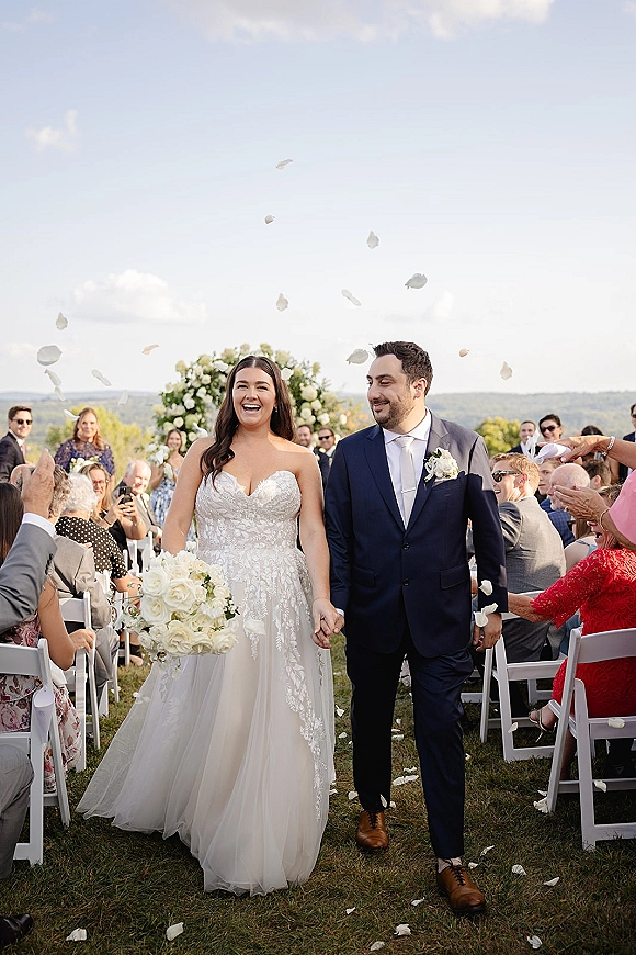 Wedding recessional as newlyweds walking down aisle holding hands, guests tossing rose petals beneath a floral arch on a grassy lawn