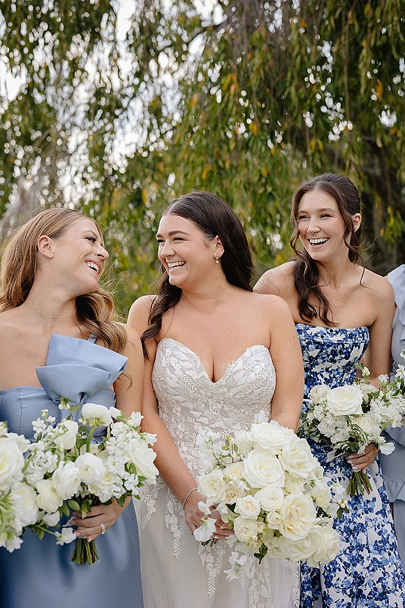 Bridesmaid group portrait with bride with bridesmaids holding white rose bouquets and greenery in a garden setting beneath trees