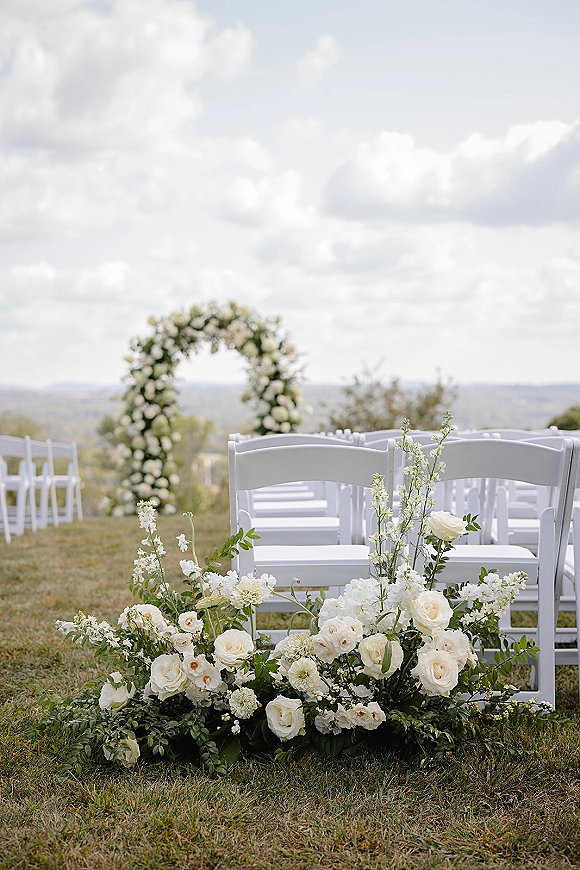 Ceremony setup for an outdoor wedding ceremony with white folding chairs facing a round floral arch of white roses on a lawn with hills