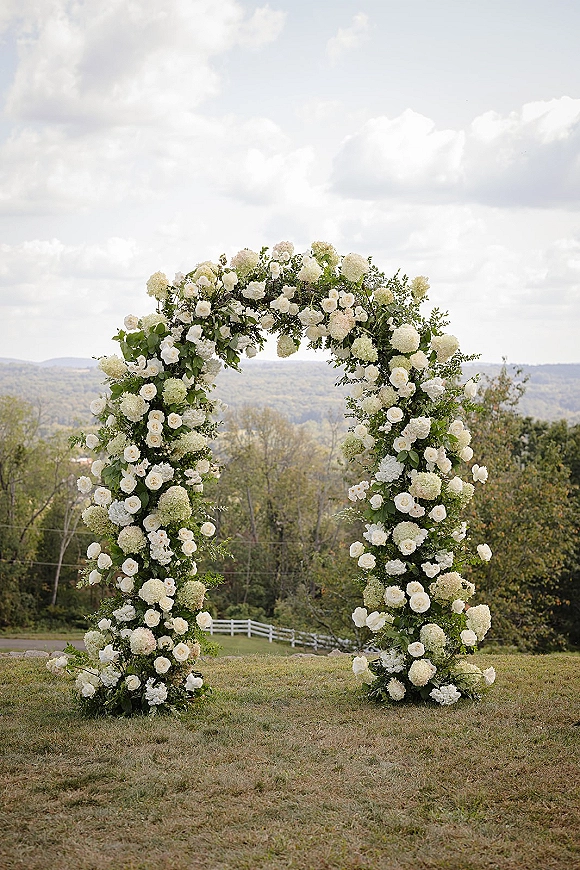 Floral wedding arch with a round floral arch design, white roses and hydrangeas with greenery on a lawn beside a white fence under clouds