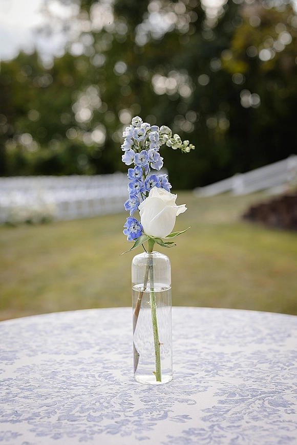 Wedding bud vase single stem centerpiece with a white rose and blue blooms in clear glass on a tablecloth, set on an outdoor lawn with ceremony chairs
