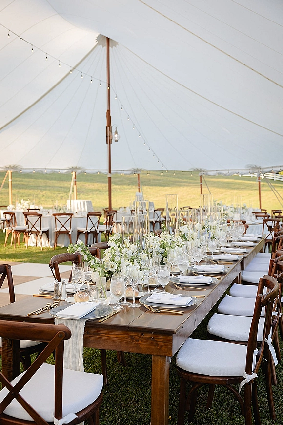 Reception tablescape on a farmhouse wedding table with gold flatware, white florals and greenery garland under string lights in a sailcloth tent