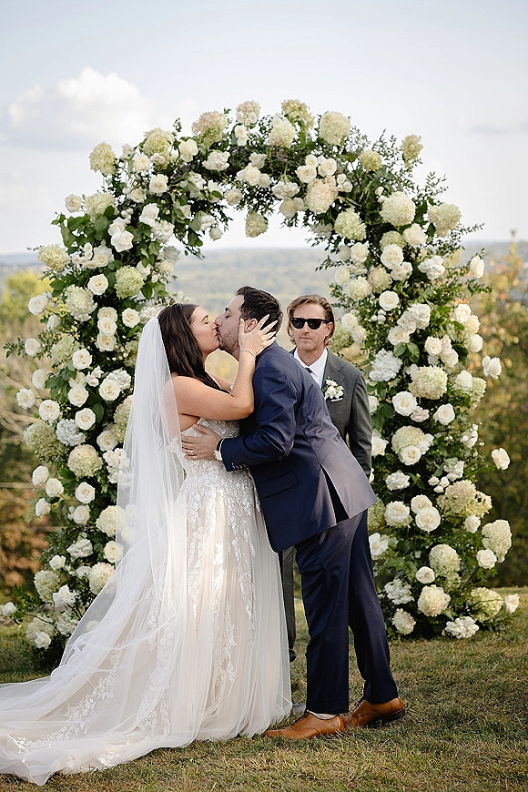 Wedding kiss moment as the bride and groom share a ceremony kiss photo under a round white rose and hydrangea arch on a hilltop lawn