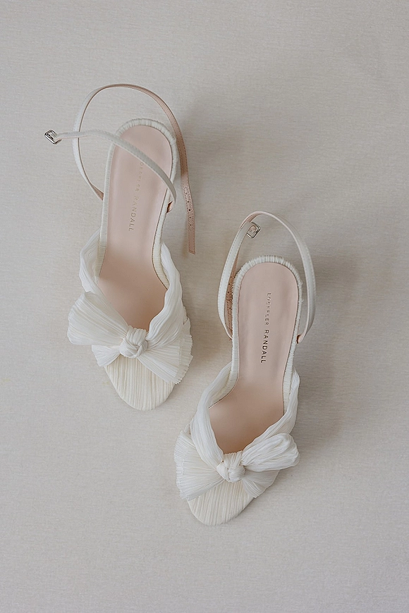 Bridal shoes, white wedding heels with pleated bow detail and slingback ankle straps resting on a neutral fabric surface flatlay