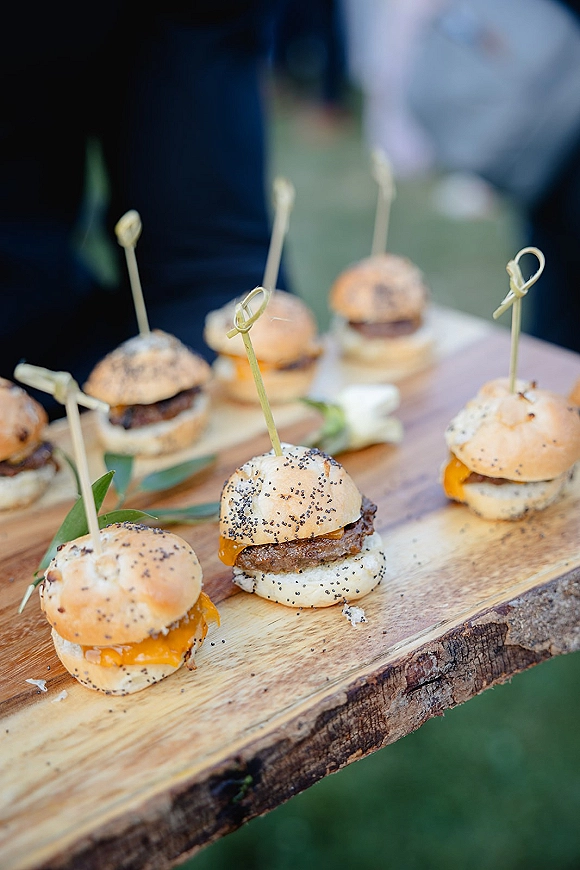 Wedding appetizers of mini slider appetizers with poppy seed buns, cheese, and bamboo picks on a wooden board, greenery garnish on an outdoor lawn