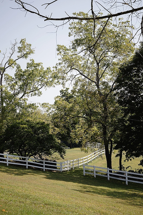 Outdoor wedding venue with a white fence bordering a green lawn, framed by trees and rolling hillside under open sky