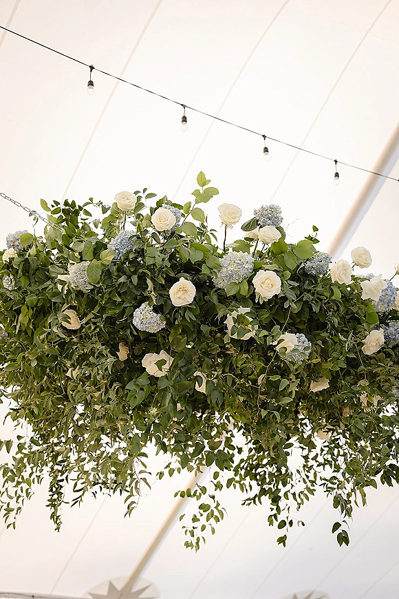 Hanging floral arrangement wedding floral chandelier with greenery, white roses, and blue hydrangeas beneath string lights on a tent ceiling