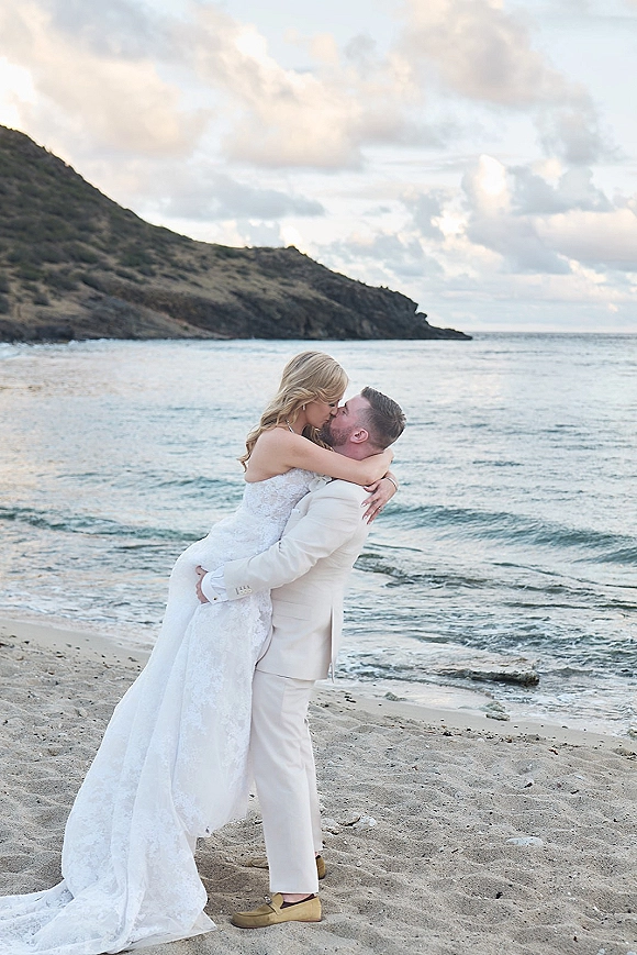 Wedding kiss portrait of the bride and groom embracing, her strapless lace dress flowing as ocean waves crash on a rocky coast under clouds