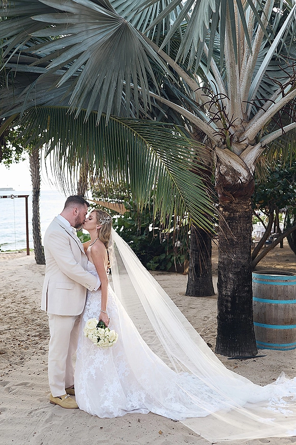 Wedding kiss portrait of bride and groom kissing, her long veil trailing in sand as she holds a bouquet with palm trees and ocean behind