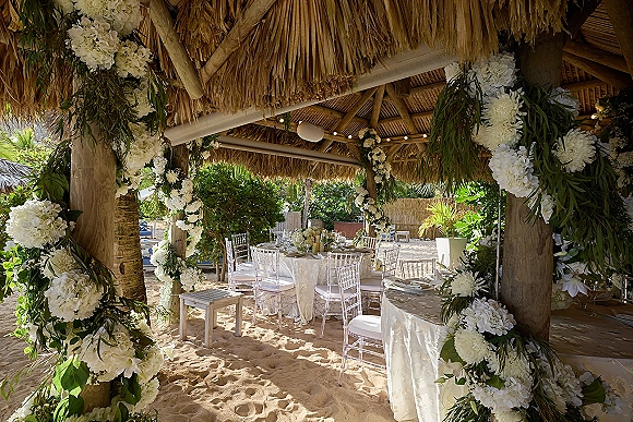 Reception tablescape for a beach wedding reception with white floral garlands, greenery, candles, and clear chairs under string lights in a thatched pavilion