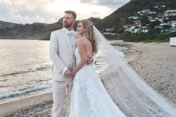 Couple portrait at a beach wedding, bride in lace strapless gown and veil blowing in wind as groom in white tuxedo embraces her by the ocean
