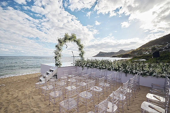 Beach ceremony setup with a white floral arch and clear acrylic chairs on sand, facing the ocean under a cloudy sky