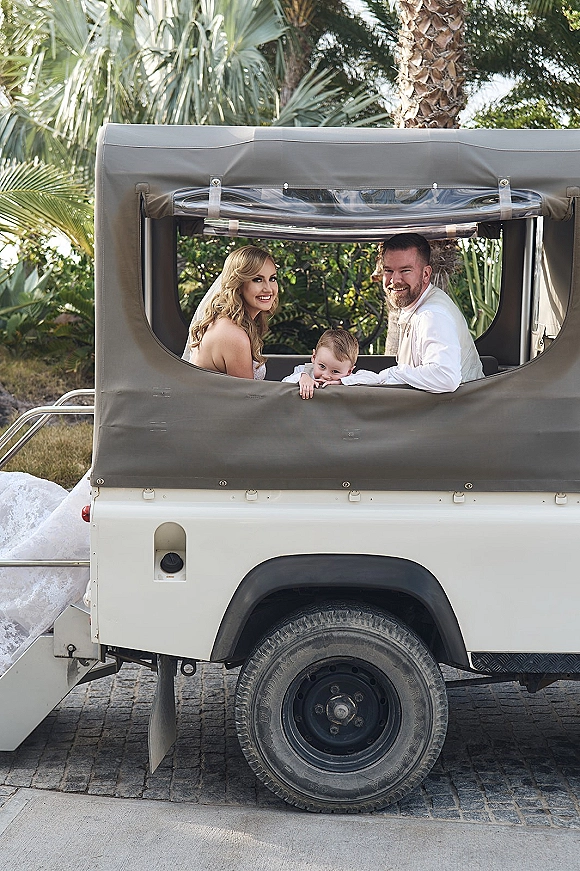 Wedding family portrait of bride groom and child in a safari jeep, bride in veil beside groom in white shirt, palm-lined driveway behind