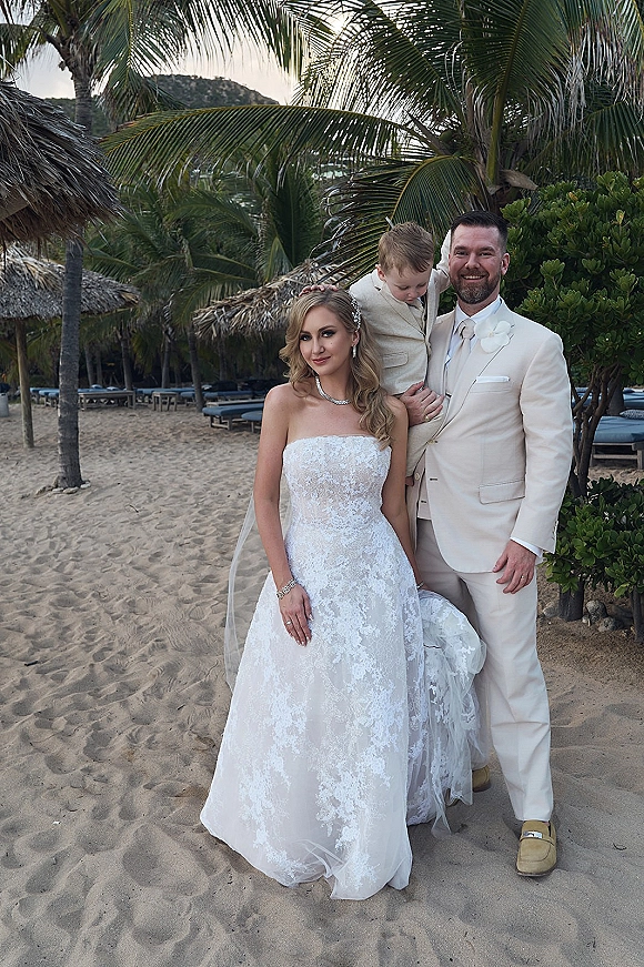 Family wedding portrait of bride in strapless lace dress and groom holding their child on a sandy beach with palm trees and palapas