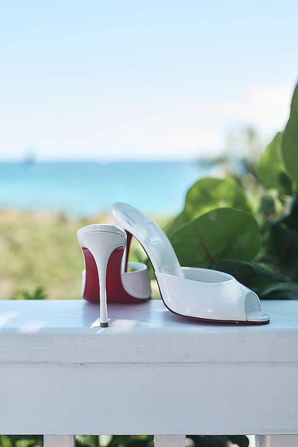 Bridal shoes, white bridal heels with red soles on a white railing, with ocean and sky behind and coastal greenery nearby