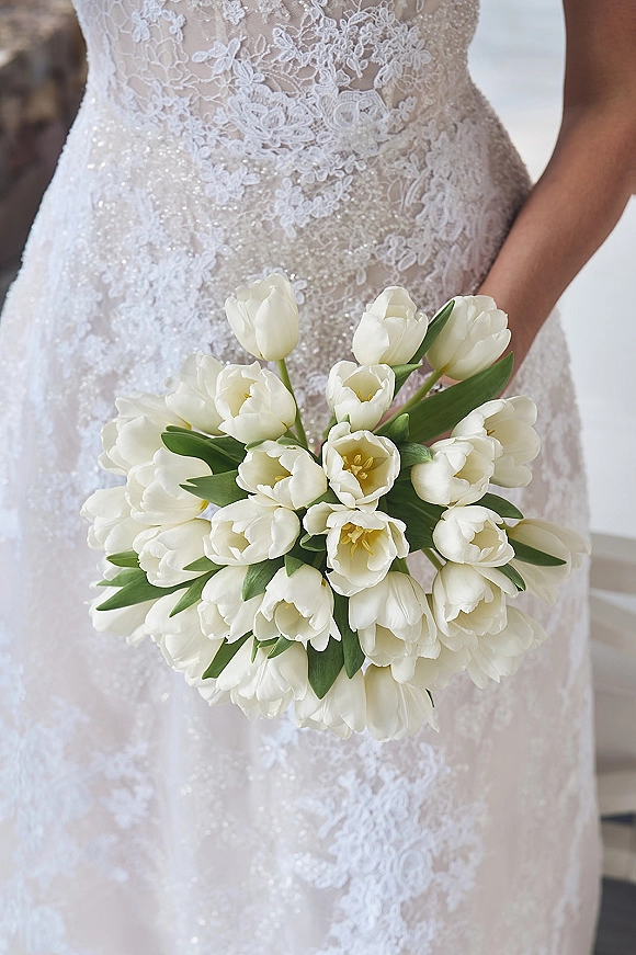 Bridal bouquet of white tulips held against a lace wedding dress, with green tulip leaves and a softly blurred outdoor backdrop