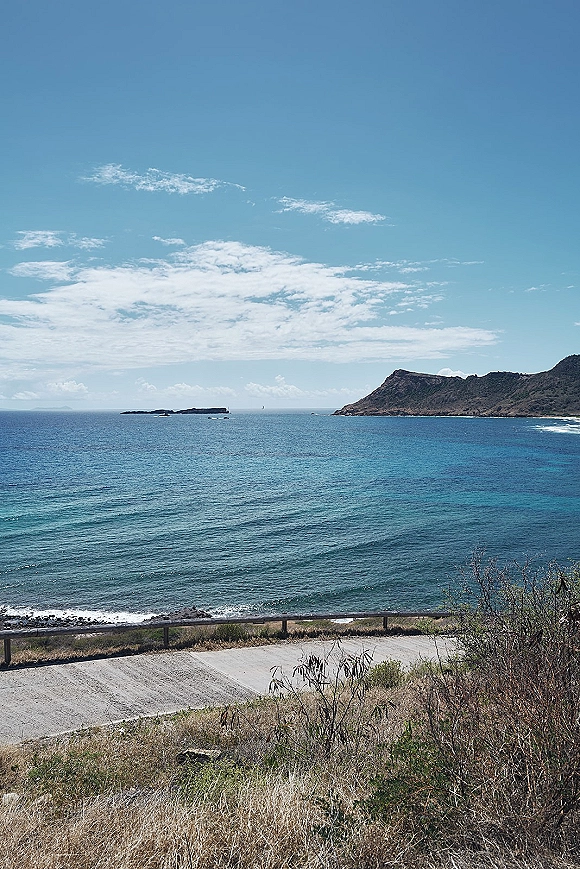 Ocean coastline coastal landscape with turquoise water and a distant island, bordered by rocky hills, coastal road, dry shrubs, and cloudy blue sky