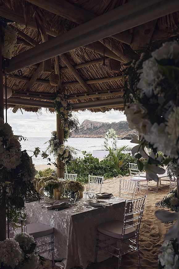 Beach wedding reception with beach reception decor under a wooden pergola, white linens, candles, florals, and oceanfront sand backdrop