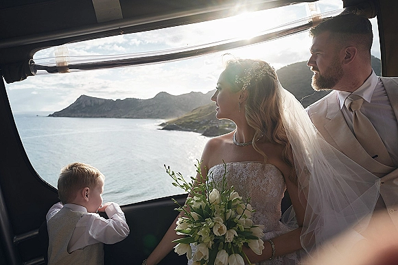 Couple portrait of bride and groom in car, bride with white tulip bouquet and veil, child in vest, ocean cliffs beyond window