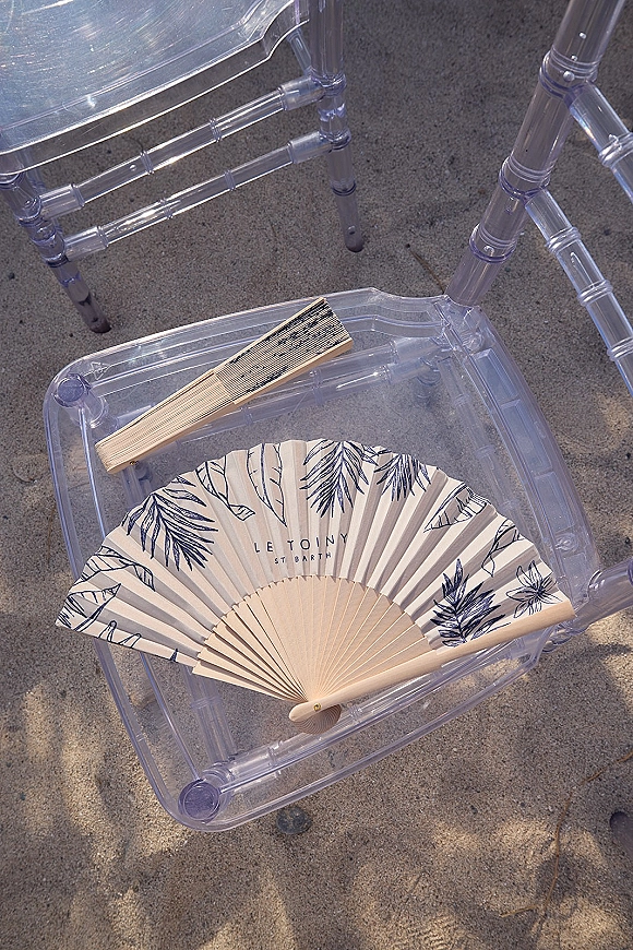 Wedding ceremony fan with wooden handle on a clear acrylic chair, personalized ceremony fans resting above sandy outdoor ground