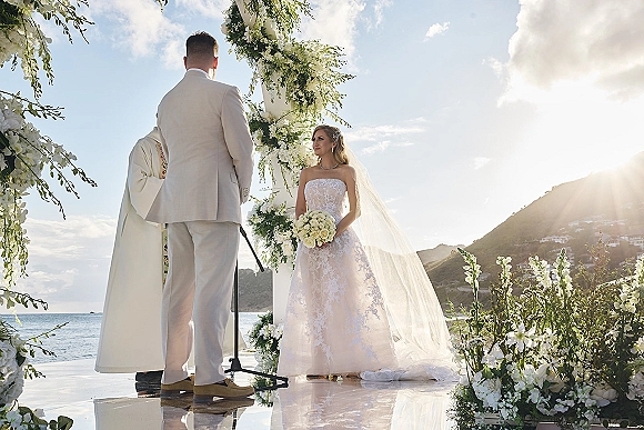 Wedding ceremony with bride and groom exchanging vows under a white floral arch, ocean coastline behind and sun flare glinting overhead