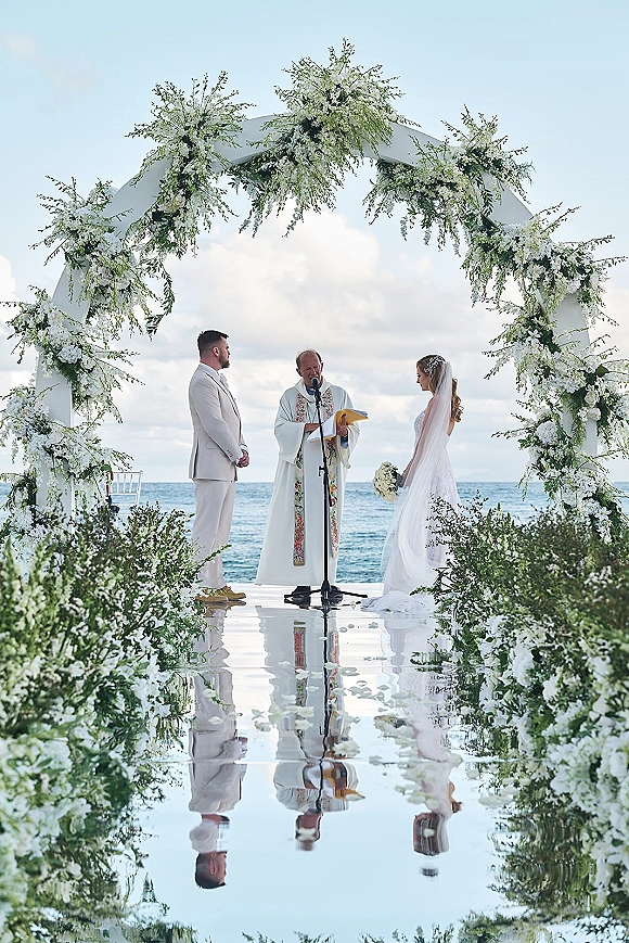 Wedding ceremony with bride and groom exchanging vows under a white floral arch, ocean horizon behind and a reflective aisle lined with florals