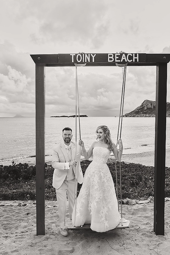 Couple portrait of bride and groom on a swing, holding ropes in wedding dress and suit beside a wooden frame sign on a cloudy beach shoreline.