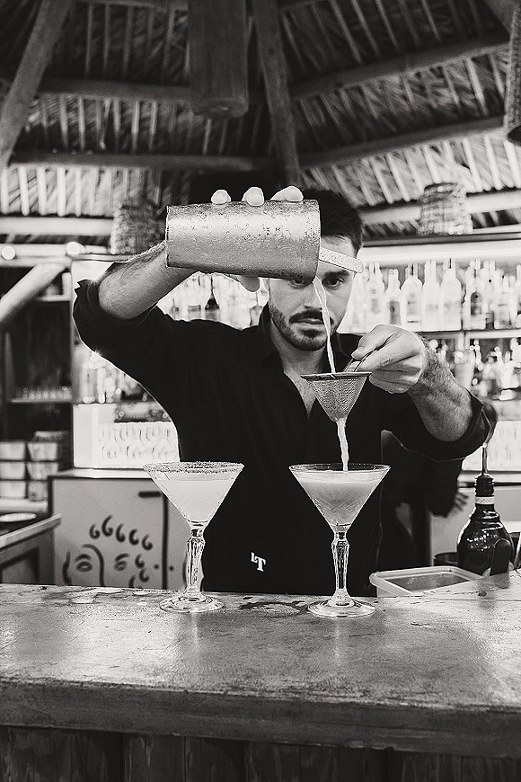 Wedding bar moment with bartender straining a cocktail into martini glasses, salt-rim accent at rustic thatched-roof bar shelves