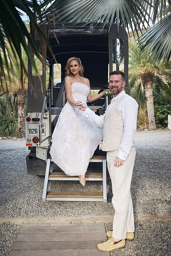 Couple portrait of bride and groom holding hands, bride in strapless lace gown beside a vintage vehicle on a palm-lined gravel drive