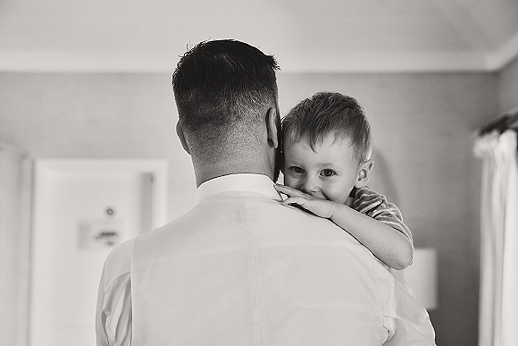 Groom with child holding his son in a dress shirt and child outfit near a doorway with window curtains in a quiet indoor room