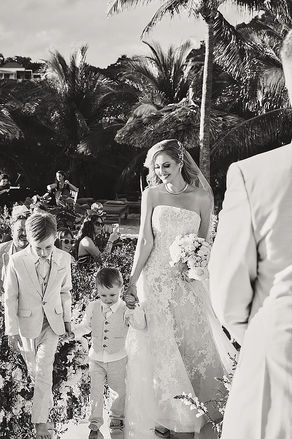 Wedding processional with bride walking down aisle holding a white rose bouquet, wearing strapless lace dress and veil in palm garden