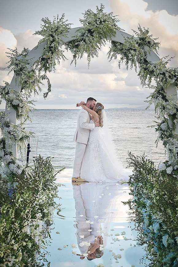 Wedding kiss at a beach wedding ceremony under a white floral arch with greenery, veil and petals on the aisle, ocean horizon behind