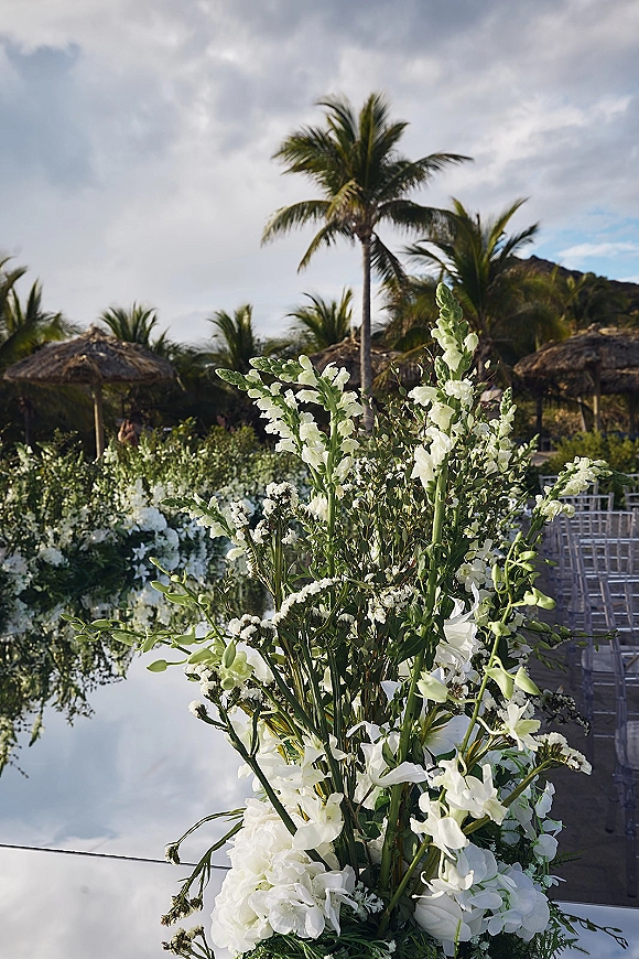 Wedding aisle decor with clear acrylic aisle panels lined by white floral arrangements and greenery, set among palm trees and thatched umbrellas