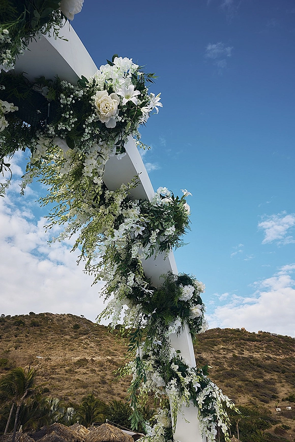 Wedding arch florals on a white floral arch with greenery garland against a blue sky with clouds, hillside, and palm trees