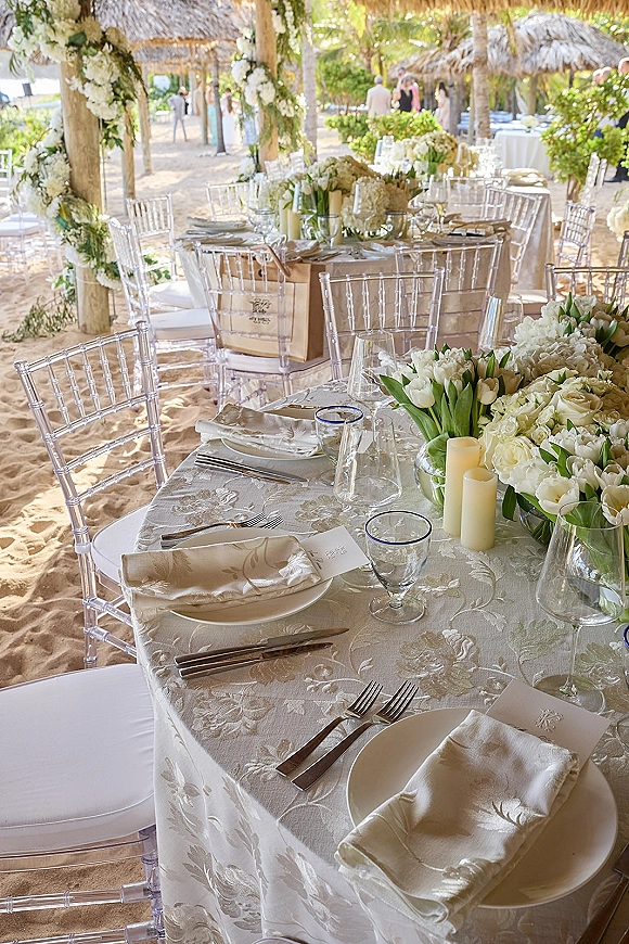 Reception tablescape with ivory damask linens, white floral centerpiece and candles, set on a sandy beach under palm trees and thatched umbrellas