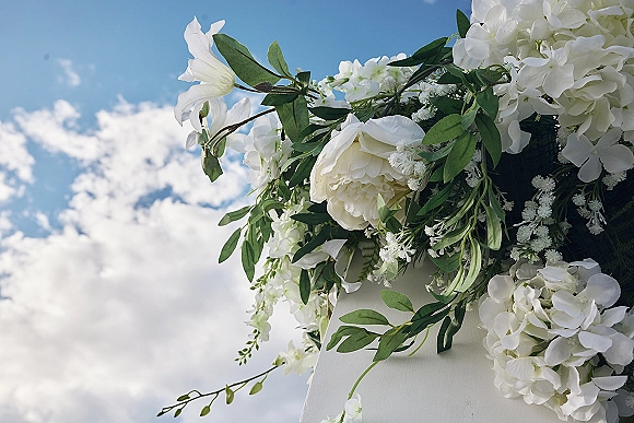 Wedding floral arch with white flower arch of peonies, hydrangeas, and greenery draping a white structure against blue sky and clouds