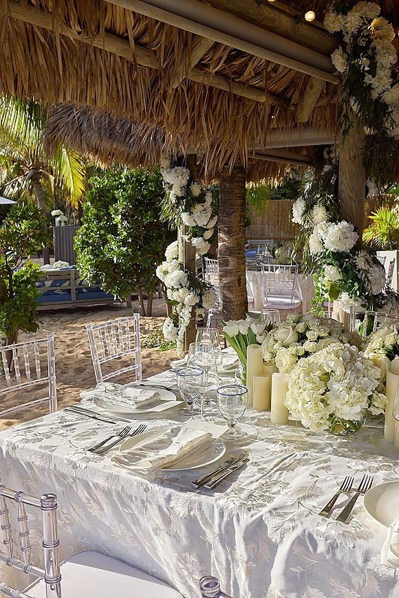 Reception tablescape with a white floral centerpiece, greenery garland and pillar candles on white linens under a thatched beach cabana with palms
