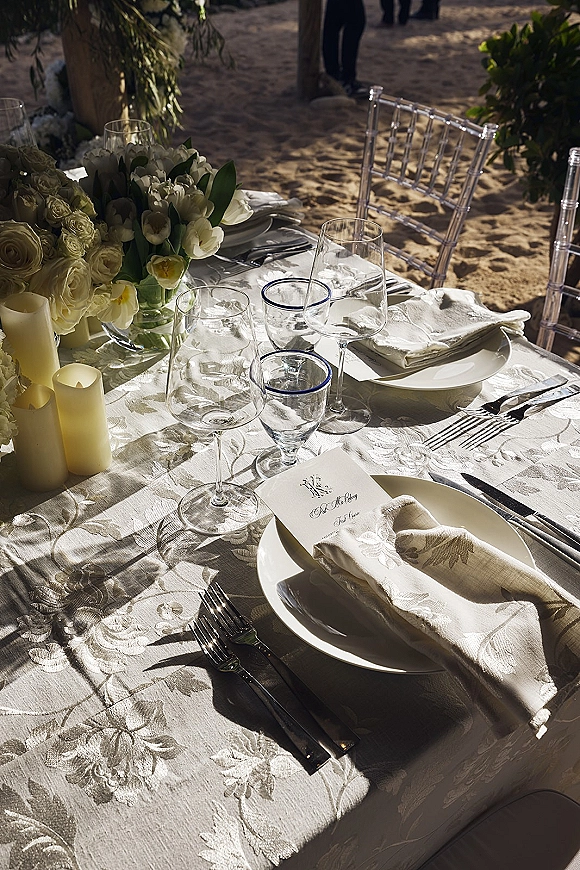 Reception tablescape with beach wedding table setting, white floral centerpiece and pillar candles on patterned cloth amid sand and greenery