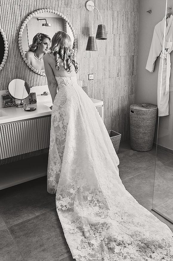 Bridal portrait of a bride at vanity mirror in a strapless lace wedding dress with long train, necklace and hair comb in a modern bathroom